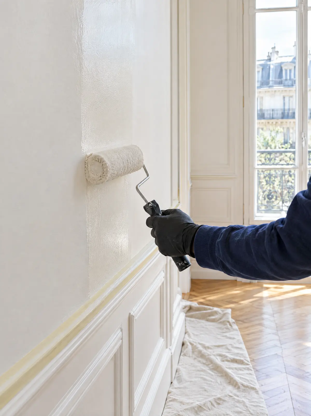 Un peintre passe le rouleau sur un mur blanc, près d’une porte-fenêtre ouverte sur la lumière.